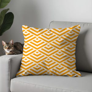 May include: Square cushion featuring a geometric design in white and orange. The cushion is placed on a grey sofa, with a cat visible on the left. The pattern is made up of repeating chevron shapes.