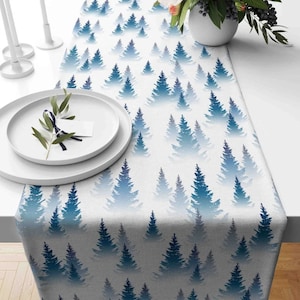 May include: A white table runner with a blue and white forest pattern. The runner features a repeating pattern of blue pine trees on a white background.