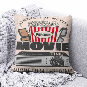 May include: A beige throw pillow with the text "Always Pop Movies Movie Time Night Film Festival" and an illustration of a bucket of popcorn, 3D glasses, and a movie projector.