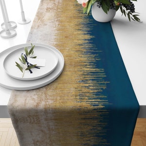 Puede incluir: Un camino de mesa azul y dorado con un patrón de rayas verticales. El camino de mesa está sobre una mesa blanca con un plato blanco y una pequeña planta verde.