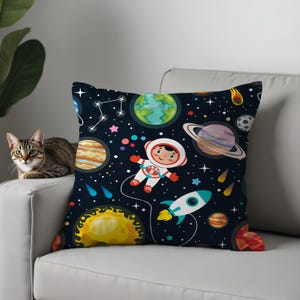 Puede incluir: Un cojín decorativo con un diseño espacial. Presenta planetas, estrellas, un cohete y un astronauta flotando en el espacio sobre un fondo azul oscuro. El cojín está sobre un sofá blanco.