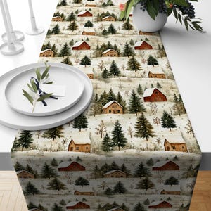 Puede incluir: Un camino de mesa con un patrón repetitivo de paisaje invernal que incluye graneros rojos, cabañas cubiertas de nieve y árboles de hoja perenne sobre un fondo color crema. Se muestra con platos y dos candelabros blancos.