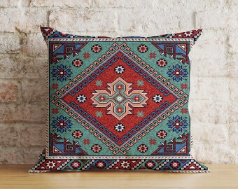 May include: Square decorative pillow with a geometric pattern in shades of red, teal, and blue. The design features a central diamond shape with a cross-like motif, surrounded by floral and geometric elements. The pillow is placed on a wooden surface.