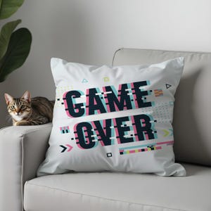 May include: White throw pillow with the words "GAME OVER" in a glitch effect design. The text is in teal, pink, and black, with geometric shapes. The pillow is on a white couch, and a cat is visible.