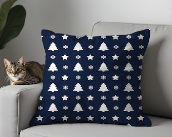 May include: A navy blue throw pillow with a repeating pattern of white Christmas trees, snowflakes, and stars. The pillow is square and rests on a white sofa. A cat is visible on the left side of the image.