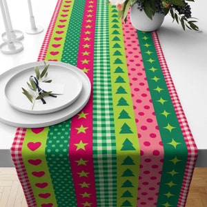 May include: A green, pink and white table runner with a repeating pattern of hearts, stars, polka dots and Christmas trees. The runner is made of fabric and has a festive holiday theme.