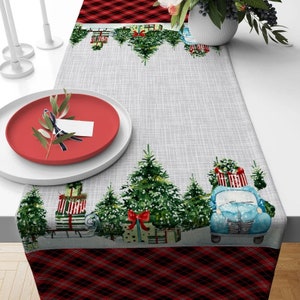 May include: A gray and white table runner with a red and black plaid border. The runner features a repeating pattern of Christmas trees, presents, and a blue vintage truck.