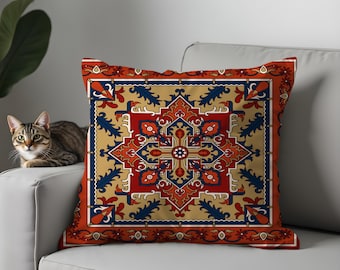May include: Square throw pillow with a detailed geometric pattern in red, blue, and gold. The pillow features an intricate design reminiscent of a traditional rug or tapestry, with a border in a darker shade of red. The pillow is on a white couch.