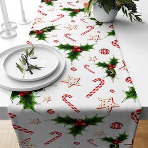 May include: White table runner with a Christmas theme featuring red and green holly leaves, red and white candy canes, and white star-shaped cookies.