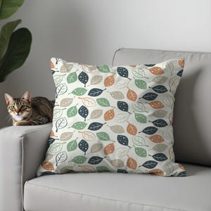 May include: A decorative throw pillow with a leaf pattern in shades of green, brown, and navy blue on a cream background. The pillow is on a gray sofa, with a tabby cat resting on the sofa arm.