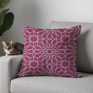 May include: A decorative throw pillow with a geometric starburst pattern in white on a deep magenta background. The pillow is square and rests on a light gray sofa. A tabby cat is sitting on the sofa.