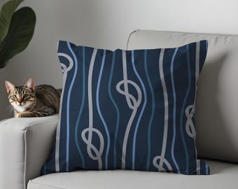 May include: Square navy blue throw pillow with a repeating pattern of wavy white and light blue lines. The lines form knots at intervals. The pillow is on a white couch, and a cat is visible in the background.