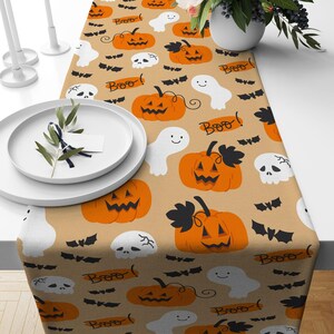 Puede incluir: Un camino de mesa con temática de Halloween con un fondo beige. El camino de mesa presenta calabazas naranjas, fantasmas blancos, murciélagos negros y calaveras blancas. La palabra "Boo!" también está impresa. Un plato y vegetación decoran el camino.