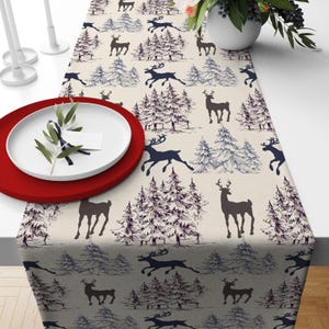 May include: A beige table runner with a repeating pattern of dark blue deer and pine trees. The trees are detailed with blue and white shading. A red plate and white plate are on the table, with a sprig of greenery.