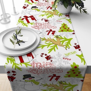 Op de afbeelding: Een witte tafelrunner met een feestelijk kerstontwerp. Het patroon bevat rode en groene pepermuntstokken, sneeuwvlokken, rendieren, kerstboom, cadeaus en de woorden "Merry Christmas".