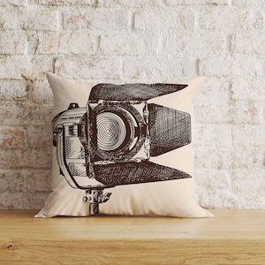 May include: A beige pillow with a black and white illustration of a movie studio spotlight.