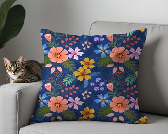 Könnte beinhalten: Ein quadratisches Zierkissen mit einem lebendigen Blumenmuster. Das Design zeigt orange, rosa, gelbe und lila Blumen sowie grüne Blätter und rote Beeren auf einem tiefblauen Hintergrund. Das Kissen liegt auf einem weißen Sofa.