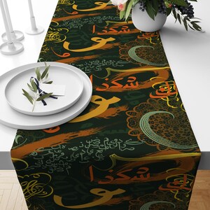 May include: A dark green table runner with a repeating pattern of Arabic calligraphy in gold, orange, and green. The calligraphy is on a dark green background with a brushstroke effect.