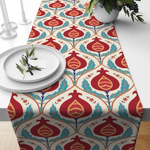 Peut inclure: Un chemin de table blanc avec un motif répété de grenades rouges, de feuilles bleues et d'accents dorés. Le chemin de table a un fond crème et une texture de lin subtile.