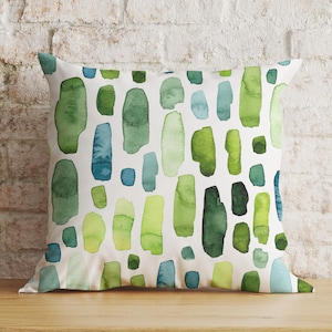 May include: A white cushion with a green and blue watercolour pattern. The pattern is made up of abstract shapes and lines.