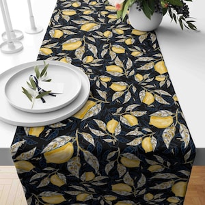 May include: A black table runner with a repeating pattern of yellow lemons and blue-green leaves. The runner is on a white table with a white plate and a sprig of greenery.