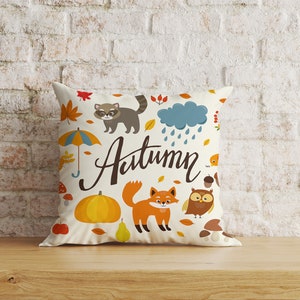 May include: A white throw pillow with a colorful autumn-themed design. The pillow features a raccoon, a fox, an owl, a pumpkin, a pear, an umbrella, and the word "Autumn".