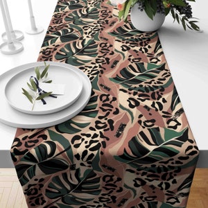 May include: A table runner with a tropical leaf and leopard print design in green, brown, and black. The runner is displayed on a white surface, with white plates and a vase of flowers in the background.