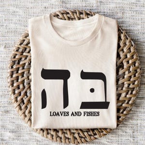 May include: A white t-shirt with a black Hebrew word and the text "Loaves and Fishes".