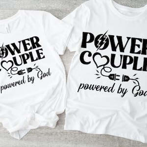 Op de afbeelding: Twee witte T-shirts met zwarte tekst en afbeeldingen. De tekst op beide shirts luidt "Power Couple powered by God". De afbeelding is een hart met een bliksem en een stekker en snoer.