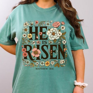 May include: Teal blue t-shirt with a floral design and the text "He is Risen" in brown lettering. The text "Matthew 28:6" is printed below the design.