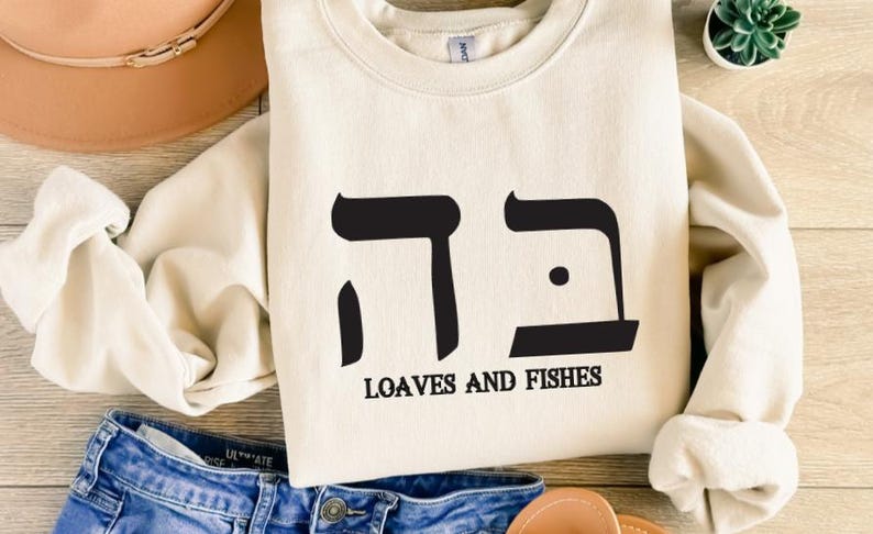 May include: A cream-colored sweatshirt with black Hebrew lettering and the text "Loaves and Fishes".