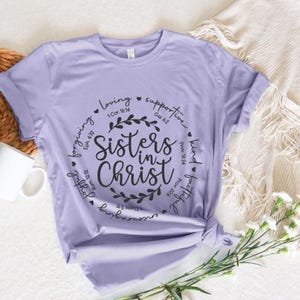 May include: A lavender t-shirt with a black circle design that says "Sisters in Christ" with bible verses and words like "loving", "supportive", "kind", and "faithful" around the circle.