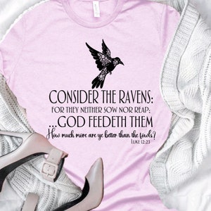 May include: A pink t-shirt with a black silhouette of a bird with floral details. The text "CONSIDER THE RAVENS: FOR THEY NEITHER SOW NOR REAP: ...GOD FEEDETH THEM How much more are ye better than the fowls? LUKE 12:23" is printed on the shirt.