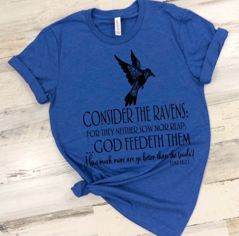 May include: A royal blue t-shirt with a black silhouette of a bird with floral details. The shirt has the text "CONSIDER THE RAVENS: FOR THEY NEITHER SOW NOR REAP: GOD FEEDETH THEM... How much more are ye better than the fowls? LUKE 12:23"