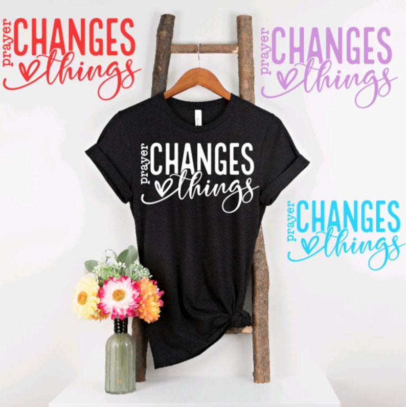 May include: A black t-shirt with the text "prayer changes things" in white lettering with a heart symbol. The text is in a cursive font. The t-shirt is hanging on a wooden ladder.