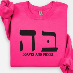May include: A pink sweatshirt with black Hebrew lettering and the words "Loaves and Fishes" printed on the front.