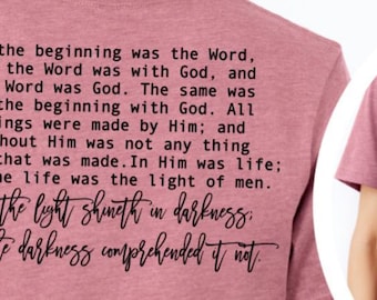 May include: Dusty rose short-sleeve t-shirt with black text on the back, including the phrase "In the beginning was the Word." The front features a small graphic of an open book with rays and the text "John 1:1-5."