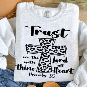 May include: A gray sweatshirt with a black and white leopard print cross design. The text "Trust in the Lord with thine Heart" is printed below the cross, along with the scripture reference "Proverbs 3:5".