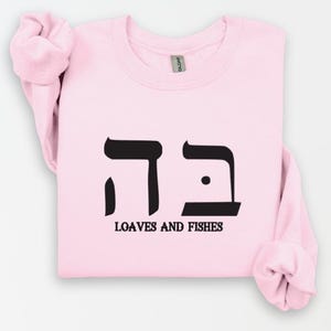 May include: Pink sweatshirt with black Hebrew lettering that says "דגים" and the words "Loaves and Fishes" underneath.