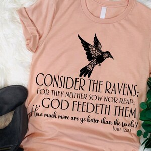 May include: A peach-colored t-shirt with a black silhouette of a bird with floral details. The shirt has a quote from the Bible, Luke 12:23, "Consider the ravens: for they neither sow nor reap; ...God feedeth them. How much more are ye better than the fowls?"