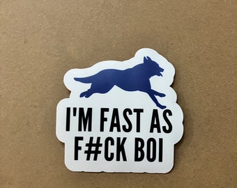 May include: A white sticker with a blue silhouette of a running dog. The sticker has the text "I'M FAST AS F#CK BOI" in black, bold letters. The sticker is on a light brown surface.
