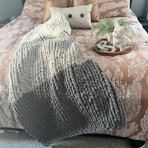 May include: A gray and white chunky knit throw blanket on a bed with a brown and white patterned duvet cover. There are two brown throw pillows and a white pillow with two black buttons on the bed. A small white ceramic dog figurine is on a wooden tray with a green plant.