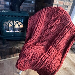 May include: A chunky knit blanket in a deep red hue, draped over a white chair. The blanket features a prominent cable knit design, adding texture and visual interest. The cozy throw is perfect for adding warmth and style to any living space.