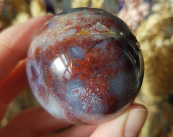 May include: A polished, red, white, and blue agate sphere with a swirling pattern. The sphere is about the size of a golf ball.