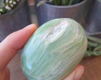 May include: A smooth, oval-shaped green stone with a layered, textured appearance. The stone is held in a person's hand, showcasing its polished surface and natural color variations. The stone's shape and color suggest it could be a decorative item or a healing crystal.