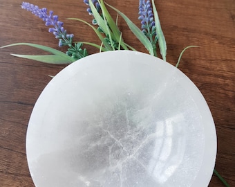 May include: A white selenite bowl with a smooth, polished surface. The bowl is round and has a slight indentation in the centre. The bowl is sitting on a wooden surface.