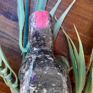 May include: A polished, dark gray stone carving with a smooth, rounded top and a flat base. The stone features a distinct, rectangular-shaped bright pink inlay at the very top, contrasting with the mottled, dark gray texture of the main body.
