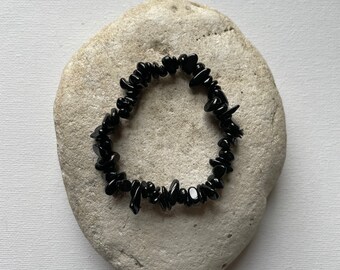 May include: A black gemstone bracelet composed of irregularly shaped, polished stones. The bracelet is resting on a light gray, smooth stone, set against a white background. The bracelet is a simple, elegant piece of jewelry.