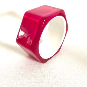 Könnte beinhalten: Ein leuchtend pinkfarbenes sechseckiges Armreif-Armband mit weißem Innenraum. Das Armband ist aus Kunststoff und hat eine glatte, glänzende Oberfläche.