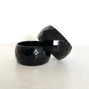 Può includere: Due braccialetti neri sfaccettati sono impilati su una superficie bianca. I braccialetti hanno una finitura lucida e un design geometrico. I gioielli sono un accessorio audace.
